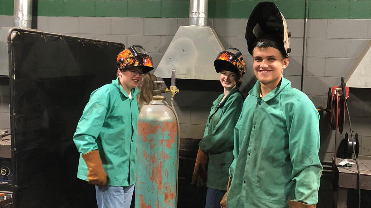 Drei Jugendliche mit Schweißhelmen und Arbeitskleidung blicken lächelnd in die Kamera