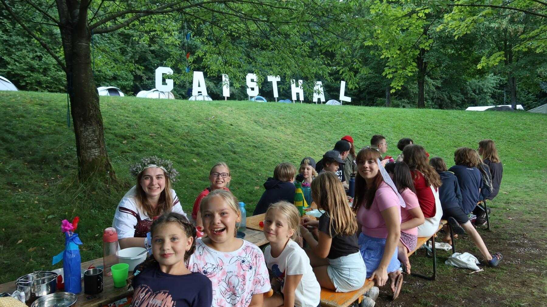 Viele Kinder sitzen mit Betreuenden auf Bierbänken auf einer Wiese