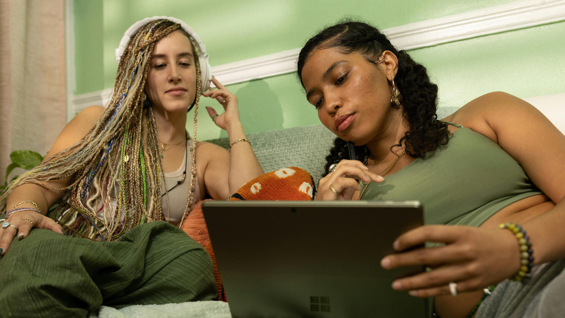 Zwei junge Frauen sitzen vor einem Tablet, eine trägt Kopfhörer, die andere hält das Gerät. Beide schauen interessiert in das Tablet.