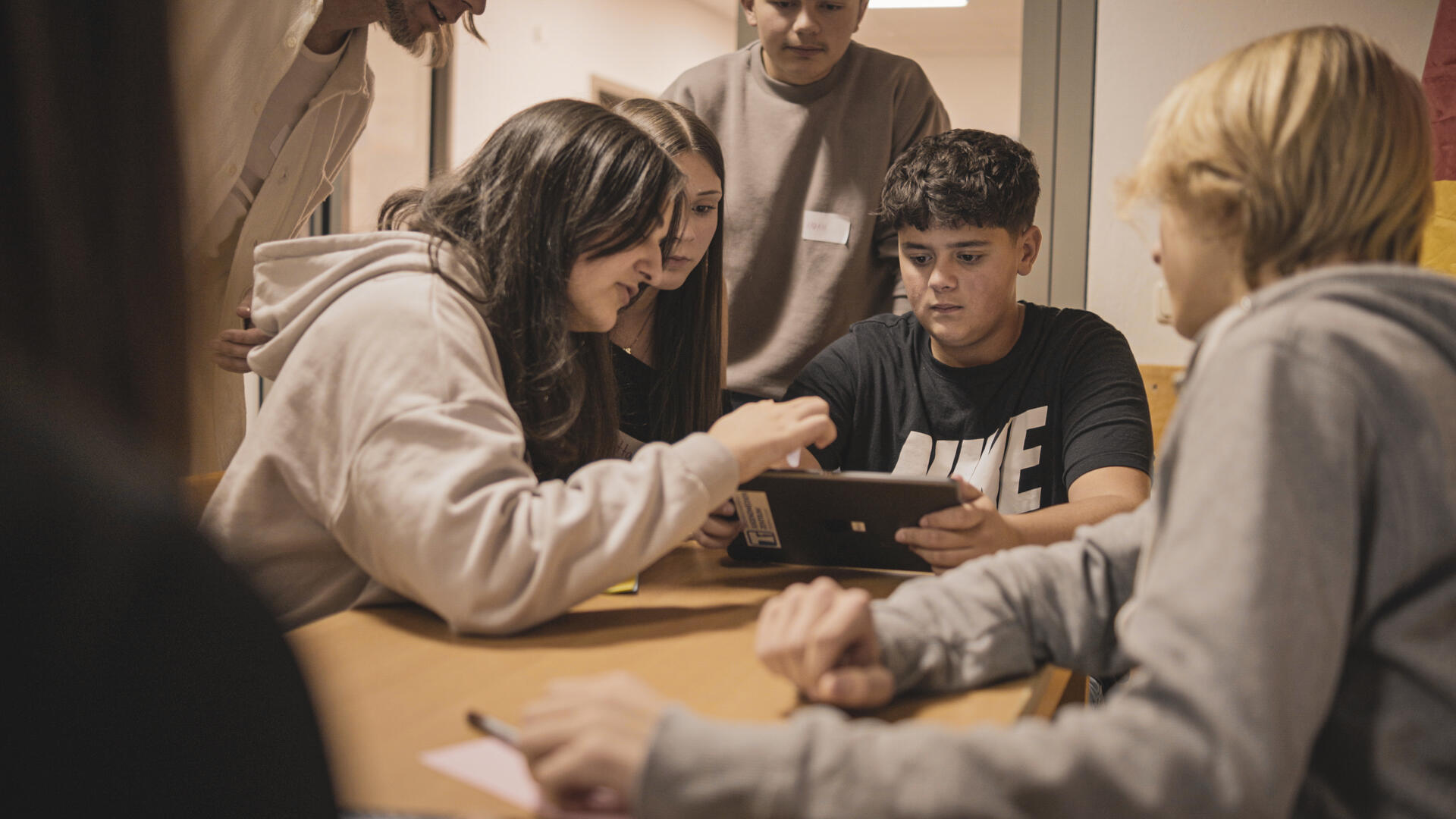 Eine Gruppe junger Menschen sitzt an einem Tisch und schaut auf ein Tablet.