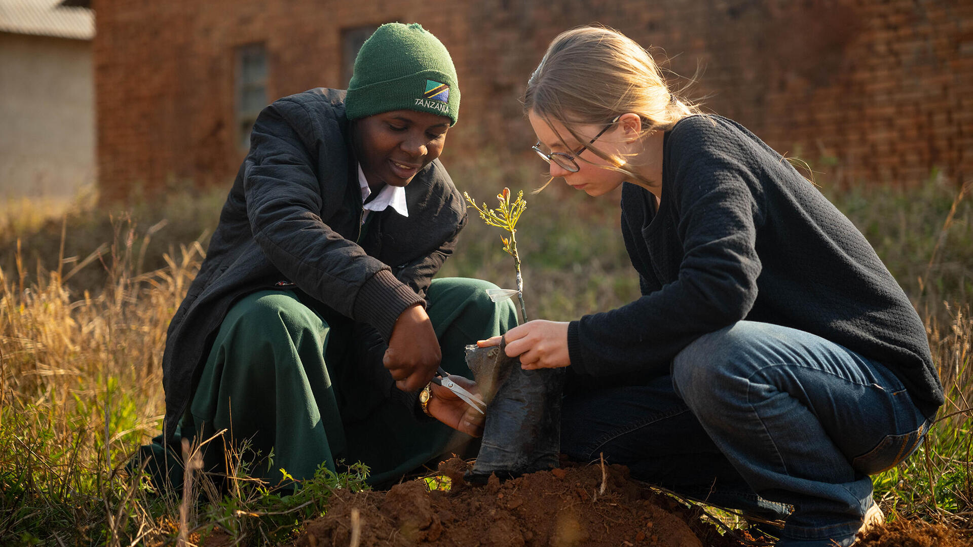 Eine tansanische und eine deutsche Schülerin setzen gemeinsam eine Pflanze in den Boden. Dazu müssen sie erst den upgecycelten Blumentopf aus alter Kleidung aufschneiden.