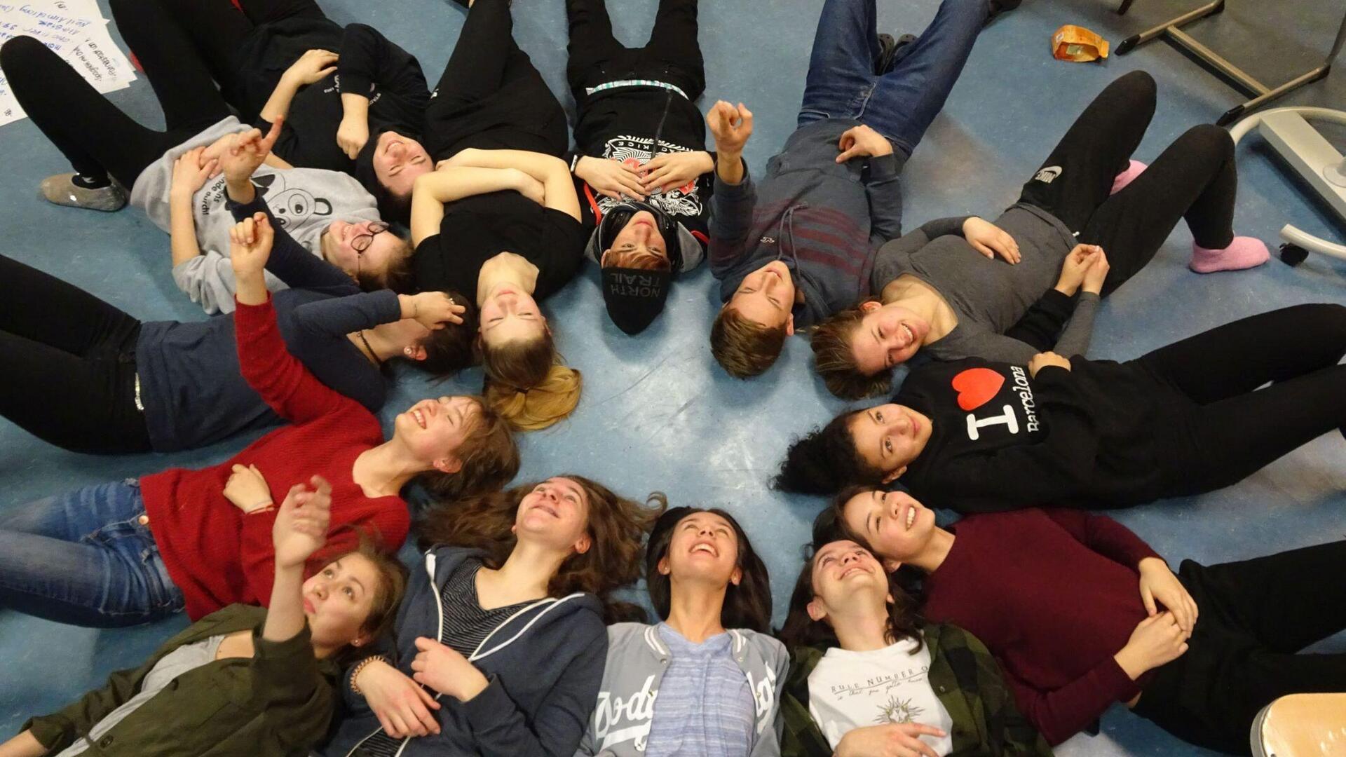 Auf dem Boden im Kreis liegende Jugendliche mit dem Kopf zur Mitte 