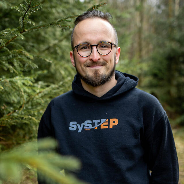 Richard Mai steht mit einem Hoodie mit der Aufschrift SyStep im Wald